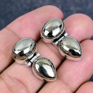 Puede incluir: Primer plano de dos pares de pendientes de pirita. Cada pendiente presenta una piedra de pirita pulida, de forma ovalada, engastada en un marco de metal plateado. Las piedras de pirita tienen un brillo metálico, reflejando la luz.