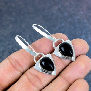 May include: A pair of silver earrings with a triangular frame and a teardrop-shaped black stone. The earrings have a hook closure and are displayed against a neutral background.