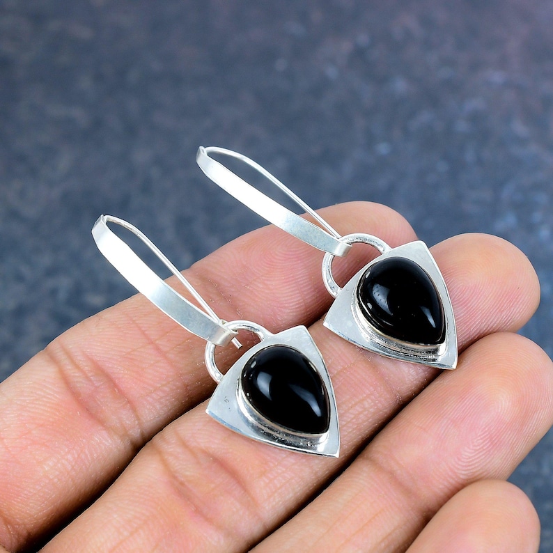 May include: A pair of silver earrings with a triangular frame and a teardrop-shaped black stone. The earrings have a hook closure and are displayed against a neutral background.