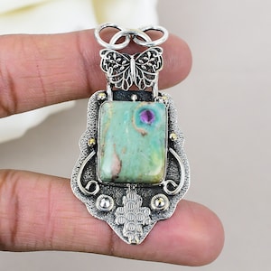 以下が含まれることがあります： 緑とピンクの宝石がフィリグリーのデザインにセットされたシルバーペンダント。ペンダントはトップに蝶のデザイン、そして詳細で装飾的なフレームを備えています。