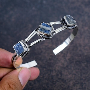 May include: A silver cuff bracelet featuring three square blue gemstones. The gemstones are set in a decorative silver frame with a twisted rope design. The bracelet has an open design and is shown against a dark background.