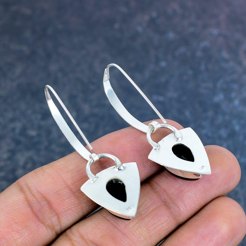 May include: A pair of silver earrings with a triangular design. Each earring features a black, teardrop-shaped stone set within a silver frame. The earrings have a hook closure.
