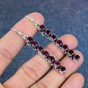 May include: A pair of silver earrings featuring a linear design. Each earring has a series of six deep red gemstones set in a silver metal. The earrings are displayed against a dark background.