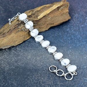 May include: A silver bracelet featuring oval-shaped white howlite stones. The stones are set in a silver metal band and connected by smaller silver links. The bracelet is displayed on a piece of wood against a dark blue background.