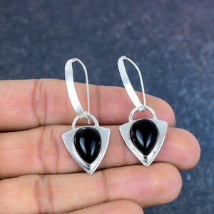 May include: A pair of silver earrings with a teardrop-shaped black stone set within a shield-shaped frame. The earrings have a long, curved hook for pierced ears. The jewelry is displayed against a neutral background.