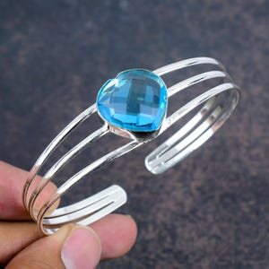 May include: A silver bangle bracelet featuring a light blue heart-shaped gemstone. The bracelet has three bands of silver.