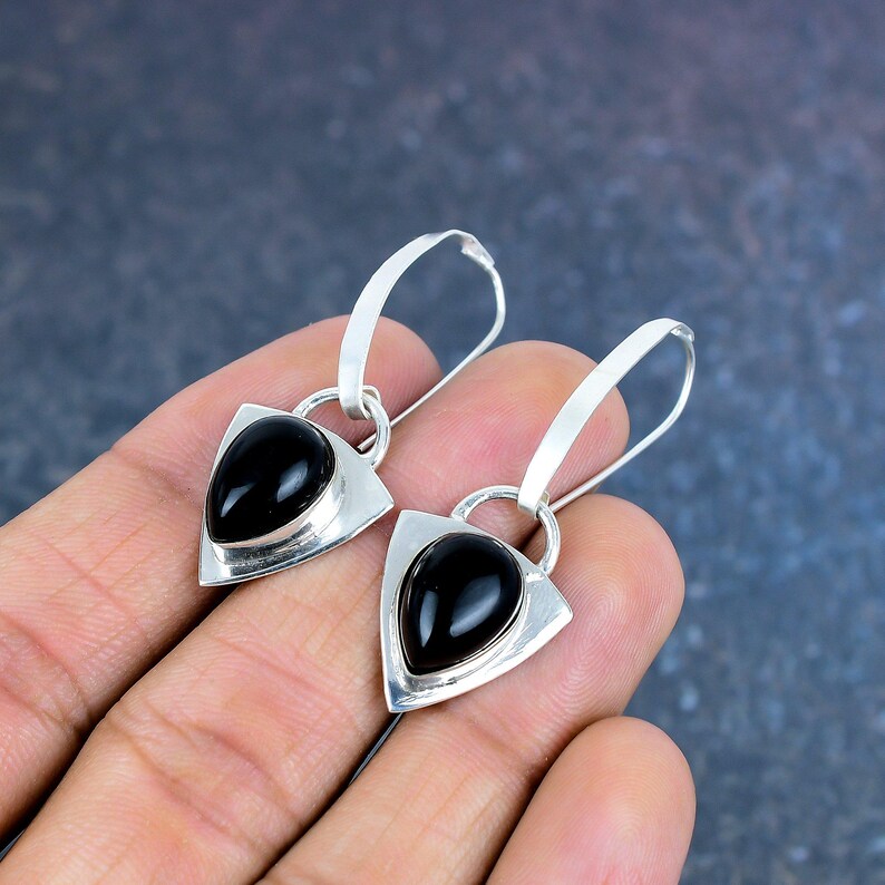 May include: A pair of silver earrings with a triangular design. Each earring features a polished black teardrop-shaped stone set within a silver frame. The earrings have a curved hook for pierced ears.