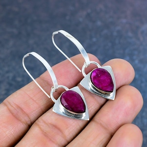 May include: A pair of silver earrings with a teardrop-shaped, deep pink gemstone set in a triangular silver frame. The earrings have a long, curved hook design.