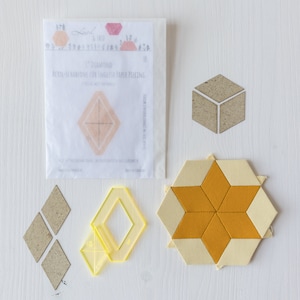 Könnte beinhalten: Eine Sammlung von Quiltzubehör, darunter rautenförmige Schablonen, Papierschablonen und ein fertiger sternförmiger Quiltblock. Die Schablonen bestehen aus Pappe und klarem gelbem Kunststoff. Der fertige Block zeigt gelben und cremefarbenen Stoff.