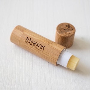 Könnte beinhalten: Eine natürliche Lippenbalsam-Tube aus Bambus mit dem Wort "NÄHWACHS" auf der Seite. Der Deckel ist abgenommen und zeigt den Balsam und das Markenlogo. Der Balsam hat eine hellgelbe Farbe.