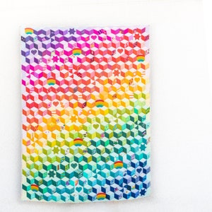 May include: A colorful quilt with a geometric cube pattern, transitioning from purple to blue, green, yellow, and red. The quilt features hearts, stars, and rainbow motifs. The quilt is hanging on a white wall.