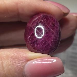 May include: A polished, oval-shaped ruby gemstone with a deep magenta-red hue. The stone features a complex internal network of natural fractures and inclusions, with a bright white circular light reflection visible on its smooth, rounded surface.