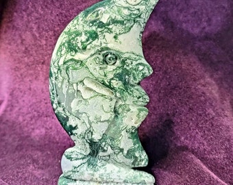 May include: Green and white carved stone sculpture of a crescent moon with a face. The sculpture is made of a type of stone with a marbled pattern.