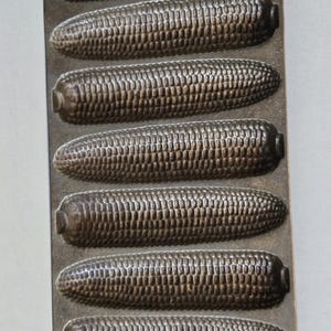 May include: A cast iron corn on the cob baking pan with seven individual molds. The pan has a handle on each end.