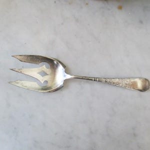 May include: A silver serving fork with three tines and a decorative handle. The fork has a pierced design with a diamond-shaped cutout. The handle is detailed with etched patterns, suggesting an antique or vintage piece.