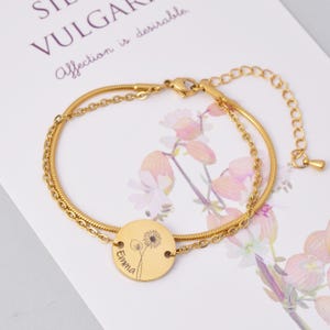 May include: Gold chain bracelet with a round charm featuring a dandelion and the name "Emma" engraved on it.
