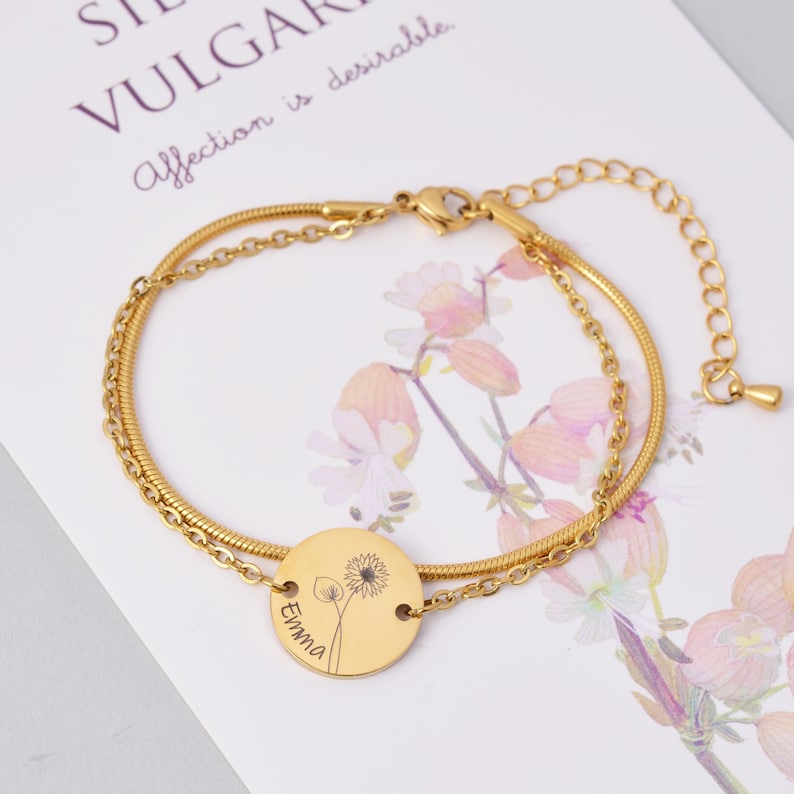 May include: Gold chain bracelet with a round charm featuring a dandelion and the name "Emma" engraved on it.