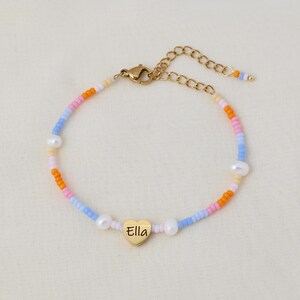 Könnte beinhalten: Ein farbenfrohes Perlenarmband mit einem goldenen Herzanhänger, auf dem "Ella" steht. Das Armband hat einen goldenen Kettenverschluss und besteht aus kleinen, bunten Perlen und weißen Perlen.