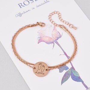 May include: Rose gold ankle bracelet with a double chain design. The bracelet has a round charm with a footprint design and the name "Leonie" engraved on it.