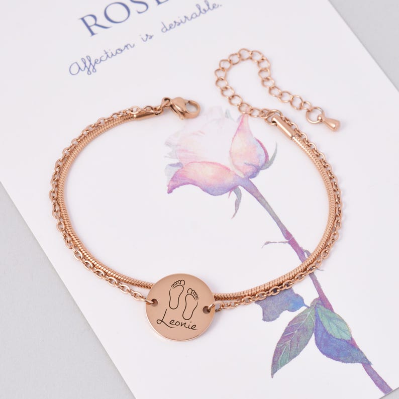 May include: Rose gold ankle bracelet with a double chain design. The bracelet has a round charm with a footprint design and the name "Leonie" engraved on it.