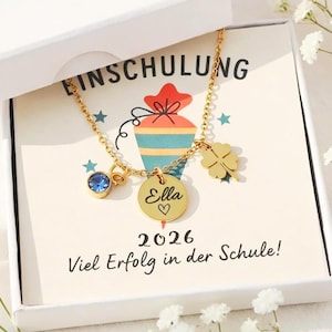 Könnte beinhalten: Eine goldfarbene Halskette mit einem blauen Edelstein, einem runden Anhänger mit der Gravur "Ella" und einem Herz sowie einem Kleeblatt-Anhänger. Die Halskette wird in einer weißen Schachtel mit einer Karte mit der Aufschrift "Einschulung" und "2026 Viel Erfolg in der Schule!" präsentiert.