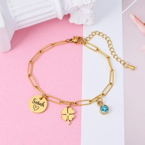 Könnte beinhalten: Goldfarbenes Armband mit einem Design aus Büroklammerketten. Das Armband hat einen runden Anhänger mit dem Namen "Sarah" und einem Herzsymbol, einen Kleeblatt-Anhänger und einen blauen Edelstein-Anhänger. Das Armband wird auf einem rosa-weißen Hintergrund präsentiert.
