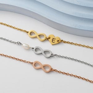 Könnte beinhalten: Drei Armbänder mit Unendlichkeitszeichen, wobei das oberste Armband einen Herzanhänger hat. Der Herzanhänger hat den Buchstaben 'E' eingraviert. Das mittlere Armband hat einen Perlenanhänger. Die Armbänder sind in Gold, Silber und Roségold.