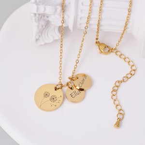 K&ouml;nnte beinhalten: Goldfarbene Halskette mit drei Anh&auml;ngern. Ein Anh&auml;nger zeigt ein Pusteblumen-Design, w&auml;hrend die anderen beiden mit den Namen "Ella" und "Clara" graviert sind. Die Halskette hat eine zarte Kette und einen Karabinerverschluss.