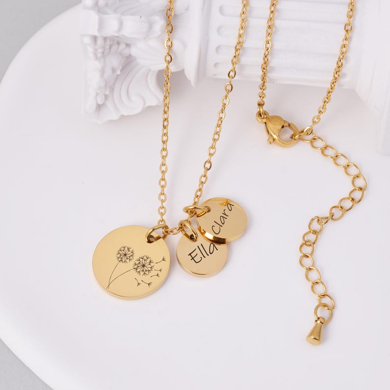 K&ouml;nnte beinhalten: Goldfarbene Halskette mit drei Anh&auml;ngern. Ein Anh&auml;nger zeigt ein Pusteblumen-Design, w&auml;hrend die anderen beiden mit den Namen "Ella" und "Clara" graviert sind. Die Halskette hat eine zarte Kette und einen Karabinerverschluss.