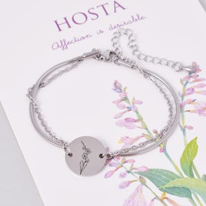 May include: A silver chain bracelet with a round charm featuring a delicate line drawing of a flower. The bracelet has a lobster clasp closure and a small extension chain.