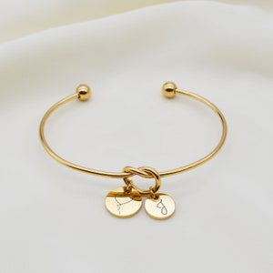 Op de afbeelding: Gouden knoop armband met twee kleine ronde bedels. De bedels zijn gegraveerd met een hart en een cursieve letter.
