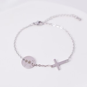 May include: A silver bracelet with a round charm featuring a dandelion design and a cross charm. The bracelet is made of a delicate chain.