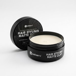 May include: A black jar of Esteem Hair Styling Matte Clay with the lid off, revealing a creamy, off-white product. The lid is next to the jar. The text on the jar reads "Hair Styling Matte Clay" and "Effortless".