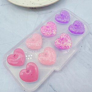 May include: A clear silicone mold with seven heart-shaped cavities filled with pink, purple, and iridescent glitter resin. The resin hearts are arranged in a gradient pattern, with the darkest purple at the top and the lightest pink at the bottom.
