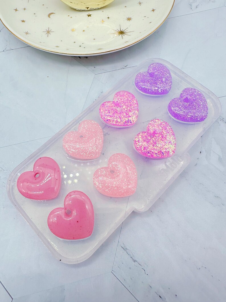 May include: A clear silicone mold with seven heart-shaped cavities filled with pink, purple, and iridescent glitter resin. The resin hearts are arranged in a gradient pattern, with the darkest purple at the top and the lightest pink at the bottom.