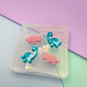 Può includere: Uno stampo in silicone trasparente con quattro forme di dinosauro. Due dei dinosauri sono rosa con glitter e due sono blu con glitter.