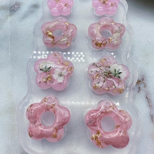 May include: A clear silicone mould with eight pink resin flower charms. The charms have a gold leaf design and some have dried flowers embedded in them.