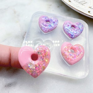 May include: A clear silicone mold with four heart-shaped resin pieces. Each heart is pink and glittery, with a hole in the center. One heart is held by a finger. The hearts are in a clear mold.
