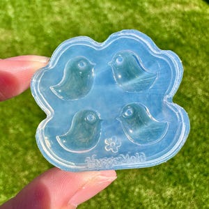 May include: A clear, light blue silicone mold featuring four bird shapes arranged in a square. The mold has a cloud-like border and a small flower design. The mold is held against a green background.