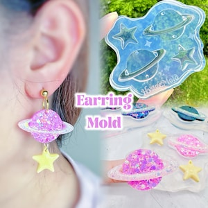 Può includere: Primo piano di un orecchino a forma di pianeta e stella con uno stampo abbinato. L'orecchino presenta un pianeta glitterato rosa con un anello bianco e una stella gialla. Lo stampo è trasparente e mostra forme di pianeti e stelle.