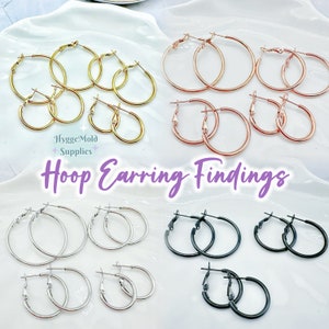 Peut inclure: Assortiment de fermoirs de boucles d'oreilles en forme de cerceau en finition or, or rose, argent et noir. Les cerceaux sont disposés en groupes de trois sur une surface blanche. Fermoirs de boucles d'oreilles en forme de cerceau.