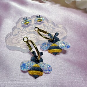 May include: A pair of gold hoop earrings with a dangling bee charm. The bee is black and yellow with iridescent glitter on its wings.