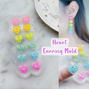 May include: A clear mould filled with colourful heart-shaped earring components in pink, yellow, green, teal, and blue. The text "Heart Earring Mold" is displayed on the image. A finished earring is shown dangling from an ear.
