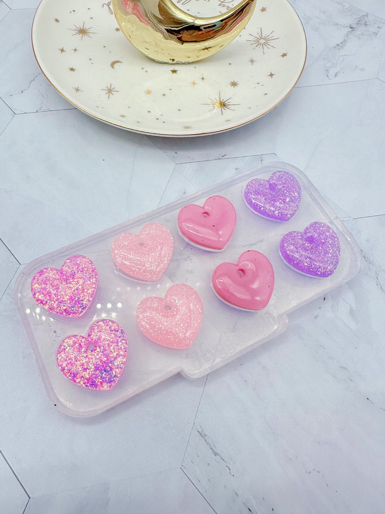 May include: A clear silicone mold with eight heart-shaped cavities. The mold is filled with pink and purple glitter hearts.