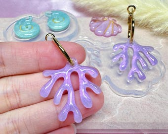 May include: A pair of lavender coral-shaped earrings with gold hoops. The earrings are held in a hand, with other seashell-shaped resin pieces and molds in the background. The earrings have a glossy finish and are a light purple color.