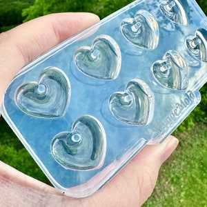 May include: Clear silicone mold with seven heart-shaped cavities. Each heart has a small hole in the center. The mold is held in a hand against a green background.
