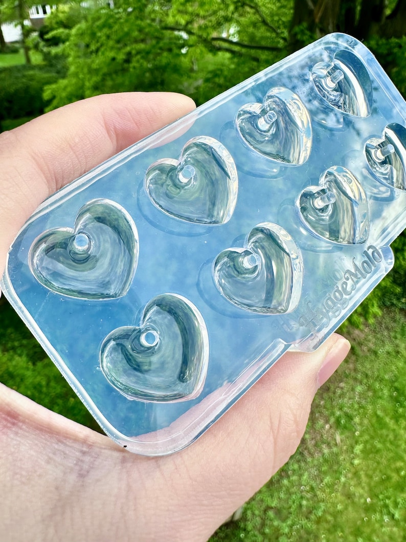 May include: Clear silicone mold with seven heart-shaped cavities. Each heart has a small hole in the center. The mold is held in a hand against a green background.