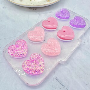 May include: A clear plastic tray with six heart-shaped resin pieces. The hearts are in shades of pink and purple and have glitter embedded in them.