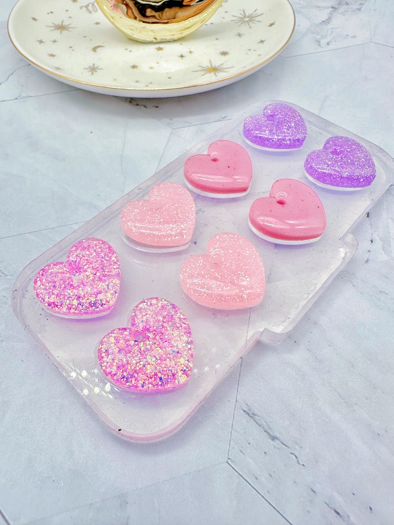 May include: A clear plastic tray with six heart-shaped resin pieces. The hearts are in shades of pink and purple and have glitter embedded in them.
