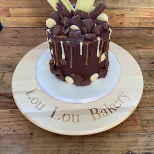 以下が含まれることがあります： 白いチョコレートとキャラメルがかけられたチョコレートケーキで、チョコレートの塊と白いチョコレートのバーで飾られています。ケーキは、「Lou Lou Bakery」と刻まれた木製ケーキスタンドの上に置かれています。